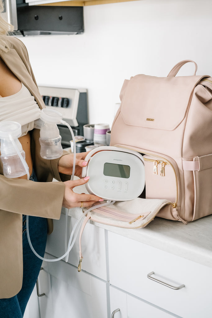 A woman setting up a breast pump next to a blush-colored vegan leather backpack with gold hardware. The bag is open, showing its spacious interior and organized compartments for storing pumping essentials