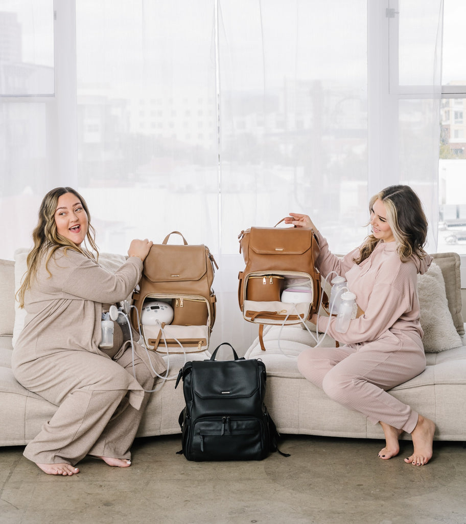 Two moms each holding up a breast pump bag. The mom on the left is holding up the Radley Pump Backpack in Toffee and the one on the right is holding up the Radley Pump Backpack in Walnut. Both have breast pumps in the bottom compartments with the Radley Cooler. This is to demonstrate that you can hold a breast pump and cooler in the bottom compartment. On the ground there is a Black Radley Breast Pump Backpack to show another color but all the compartments are closed.