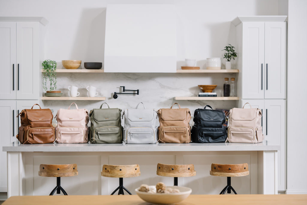 Seven vegan leather breast pump backpacks in various colors (toffee, blush, olive, mist, walnut, black, and stone) neatly displayed on a kitchen countertop, with a modern kitchen setting in the background featuring wooden shelves, bowls, and potted plants.
