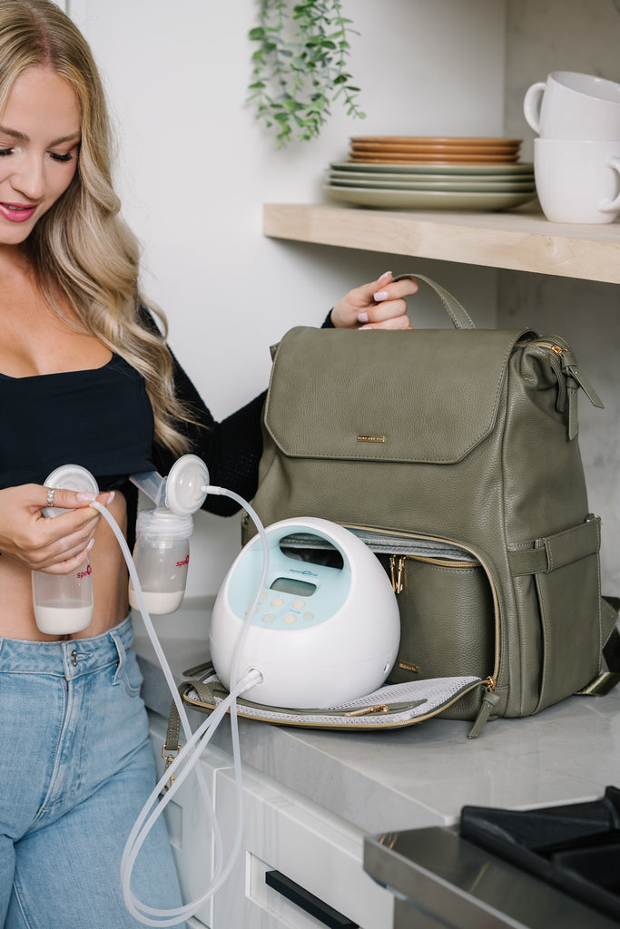 Model has her hand on her breast pump. She is wearing a black Amelia breast pump cami by Davin and Adley. She is using the Radley Breast PumP Backpack in Olive. There is a Radley Breastmilk Cooler in the bottom compartment accompanied by the Blue Spectra Breast Pump Battery. The mom is in a clean minimal kitchen and is pumping.