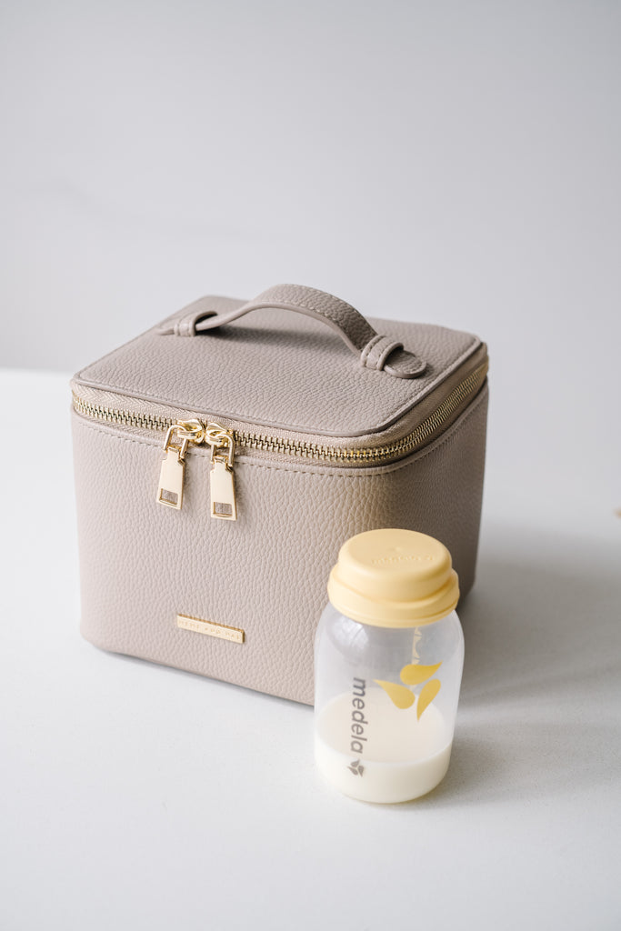 A stone-colored vegan leather breast milk cooler bag with gold hardware and a sturdy top handle, displayed alongside a Medela bottle filled with milk, showcasing a stylish and functional solution for breastfeeding moms.