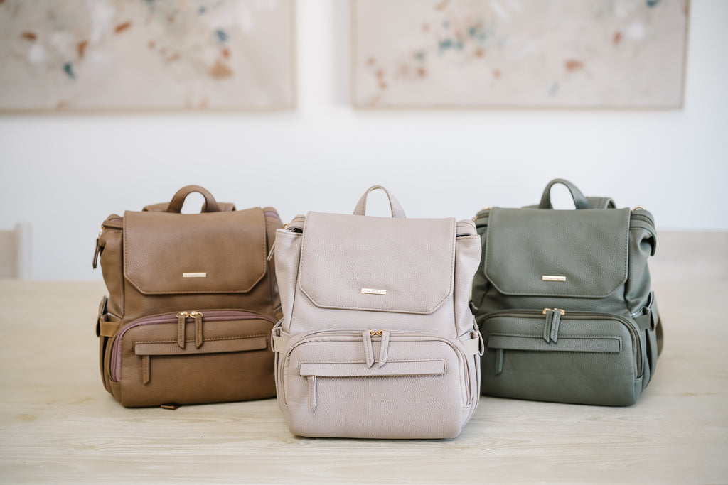 Three stylish vegan leather breast pump backpacks displayed on a light wooden table in toffee, stone, and olive colors. Each backpack features a sleek, structured design with gold accents and multiple compartments for organization.
