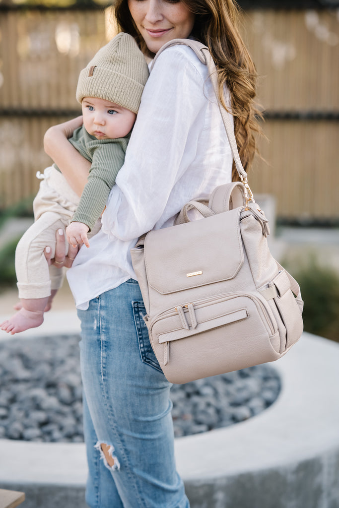 A mother holding her baby while wearing a stone-colored vegan leather breast pump backpack with gold hardware, showcasing its stylish and practical design for moms on the go.